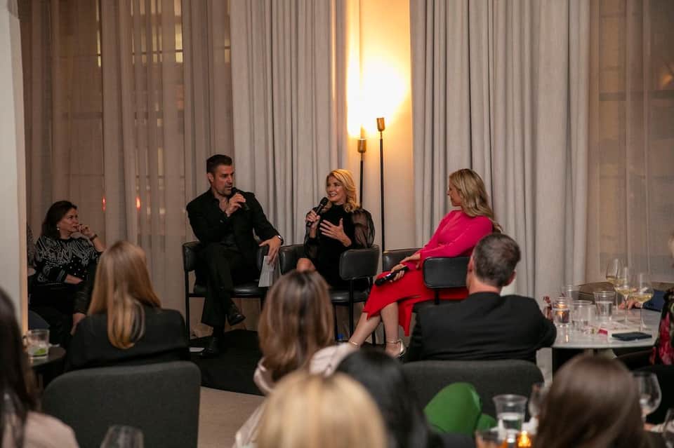 Panel discussion with four speakers seated on stage before an audience in an elegant event space with floor-to-ceiling curtains