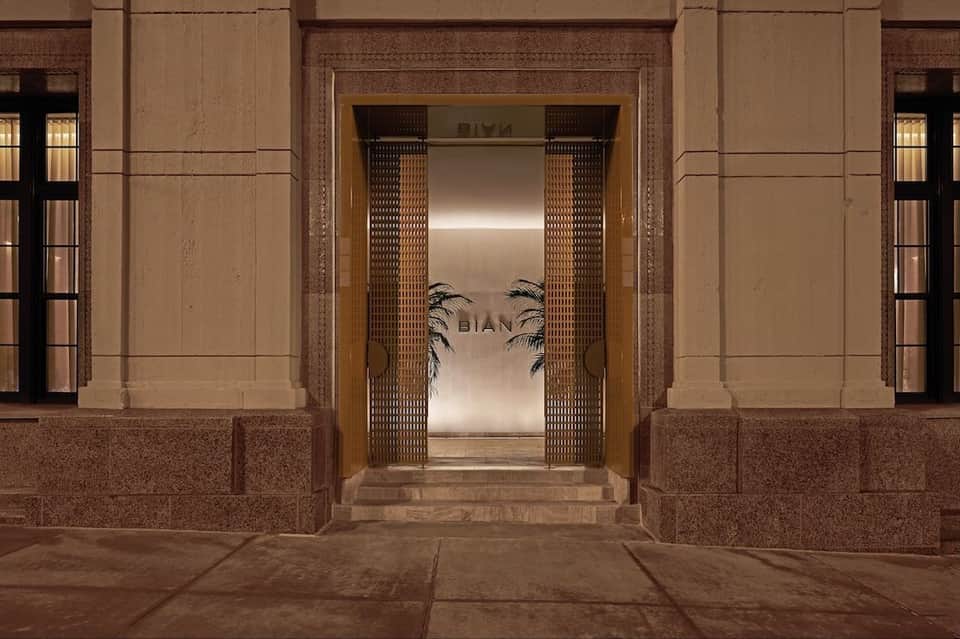 Elegant entrance to BIAN with ornate wooden doors, brass detailing, and neutral stone architectural framing
