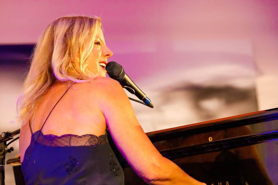 Woman with blonde hair singing at a microphone wearing a purple sequined dress, illuminated by pink stage lighting