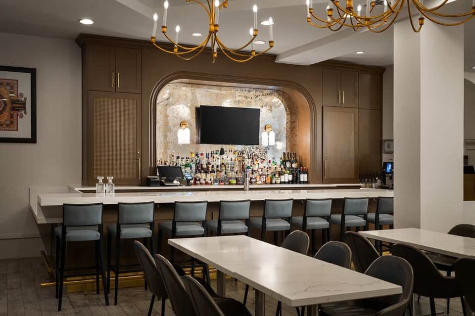 Elegant hotel bar with brown cabinetry, gold chandelier, white marble counter, and gray upholstered bar seating