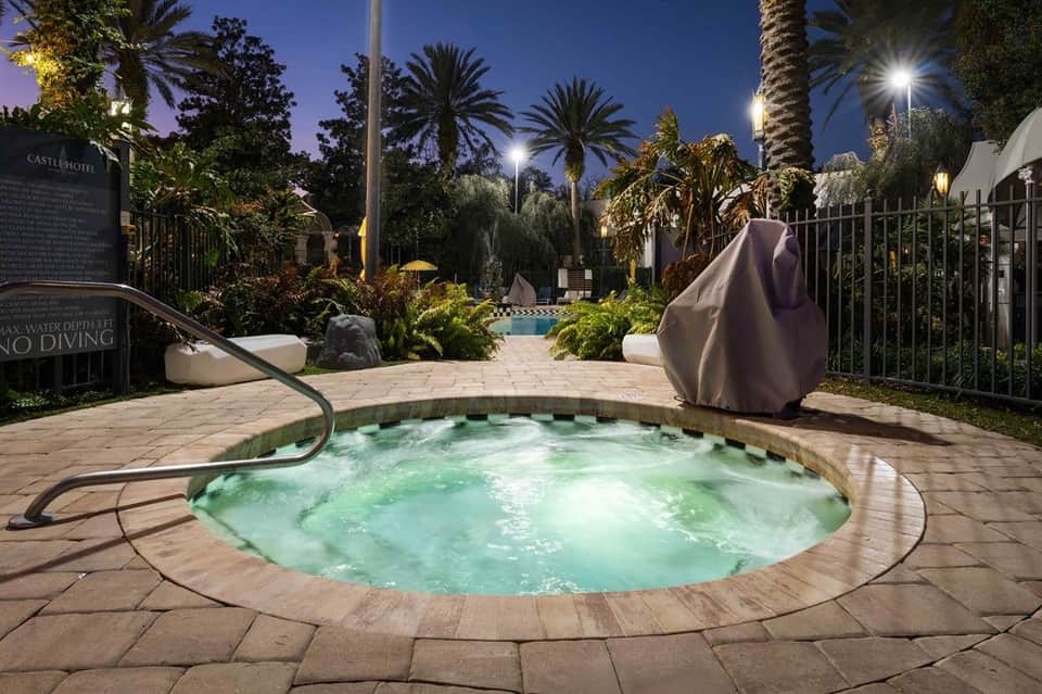 Illuminated spa hot tub at night with palm trees, landscaping, and covered lounge chairs nearby