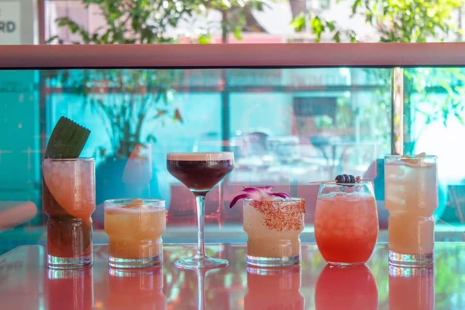 Six colorful cocktails displayed on a bar counter with turquoise windows and greenery in background