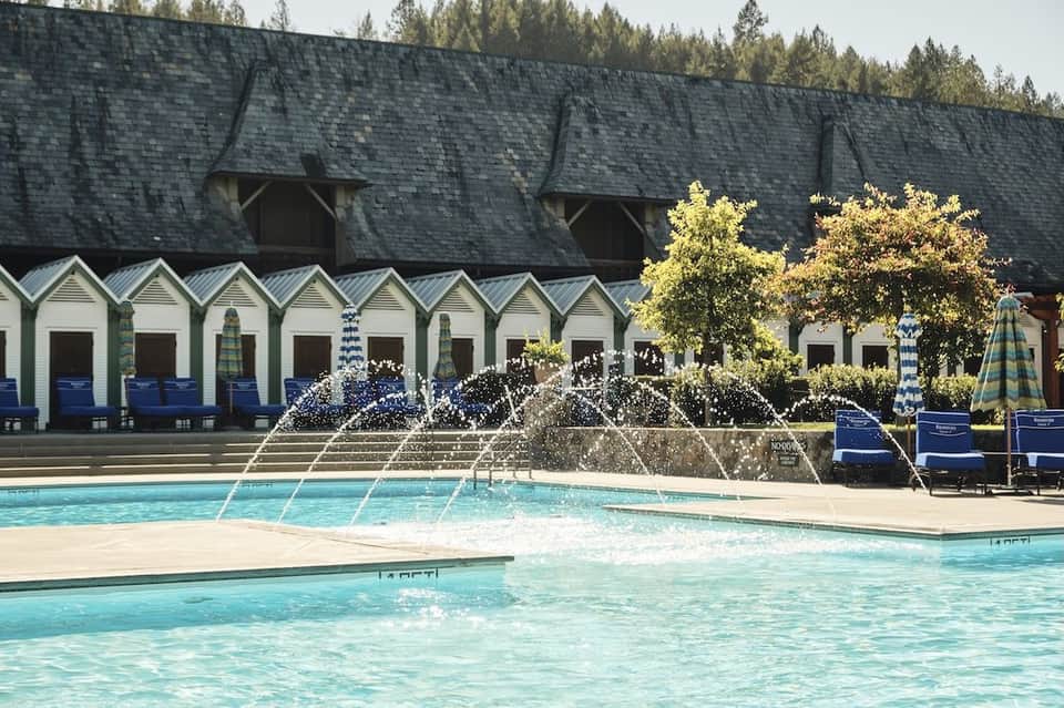 Swimming pool with water fountains and white beach cabanas backed by dark-roofed building