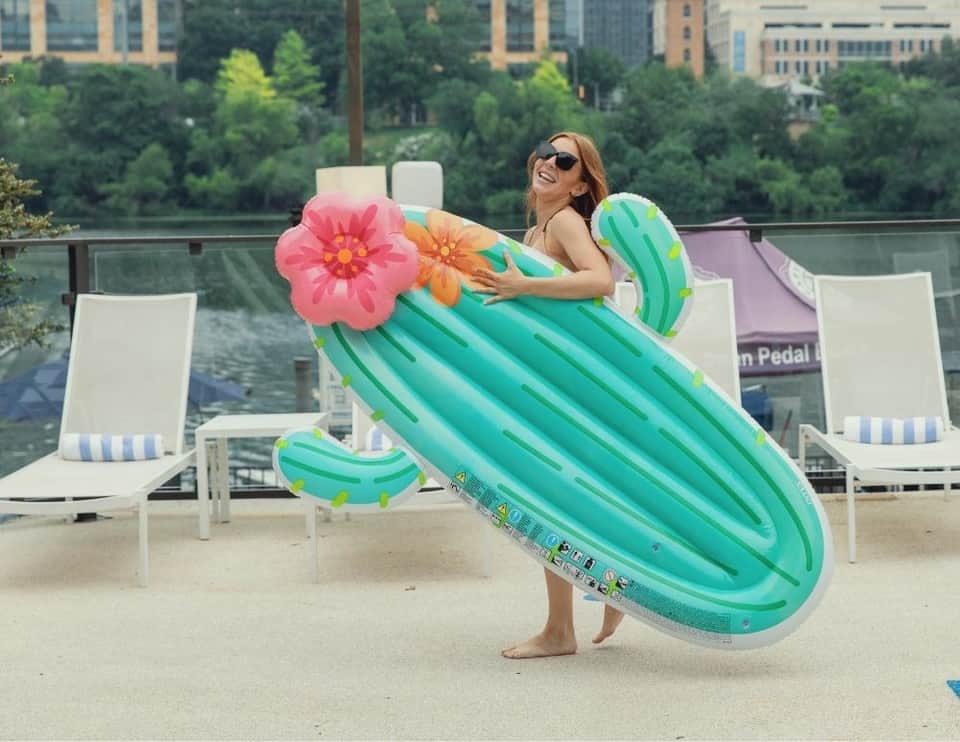 Woman holding inflatable cactus float by waterfront pool with city views