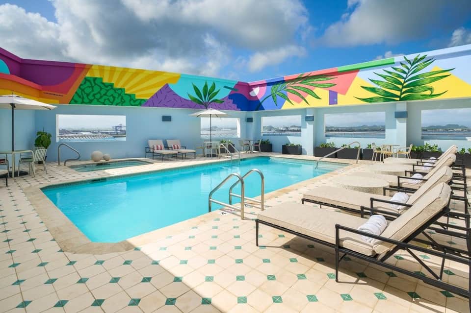 Rooftop indoor pool with colorful geometric ceiling mural and waterfront views, lounge seating