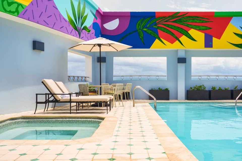 Hotel pool deck with vibrant abstract mural ceiling, lounge seating, hot tub, and ocean views