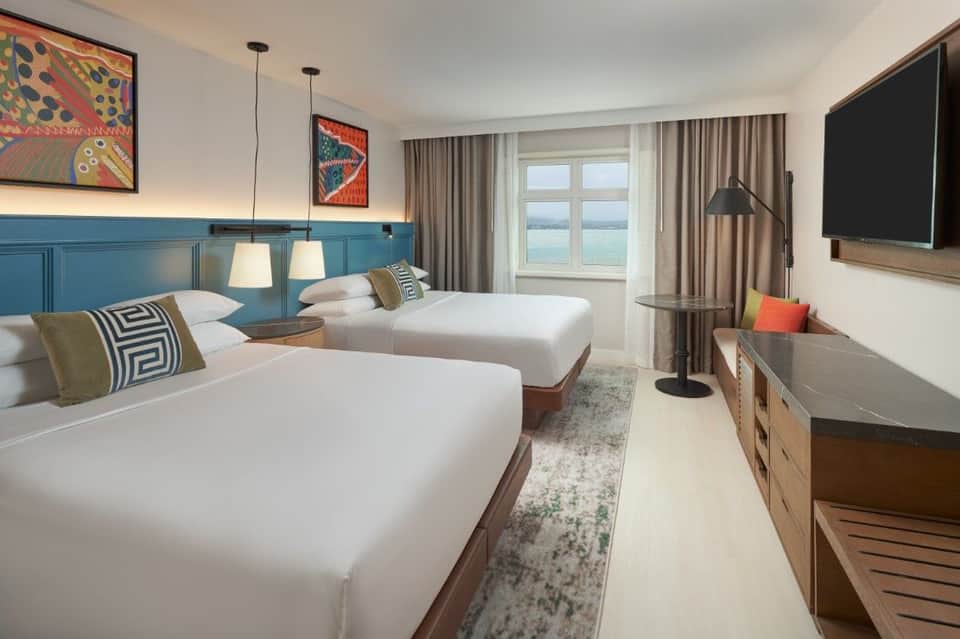 Hotel room with two queen beds, blue paneled headboard, pendant lighting, and water views