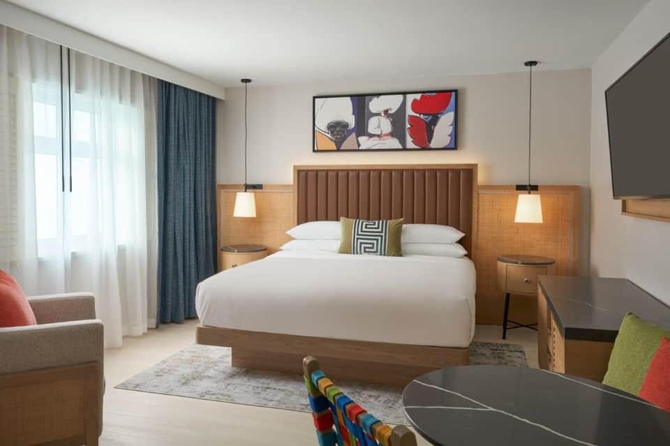 Contemporary hotel bedroom with wood-framed bed, pendant lighting, flat-screen TV, and seating area
