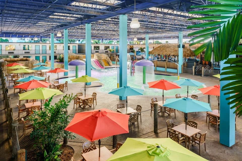 Indoor water park facility with pools, colorful umbrellas, tropical plants, and wave pool