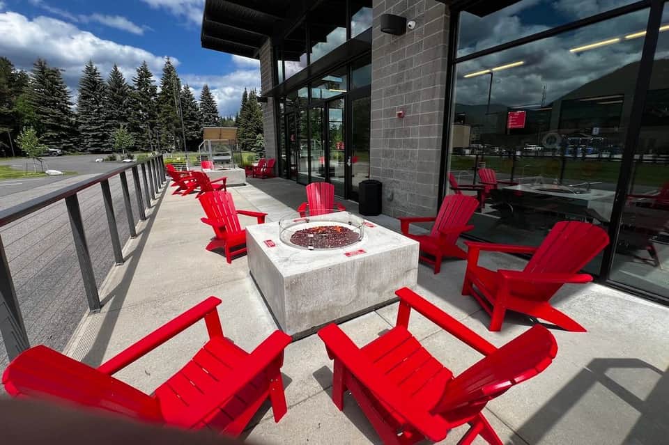 Outdoor patio lounge area with fire pit, red Adirondack chairs, and forest backdrop