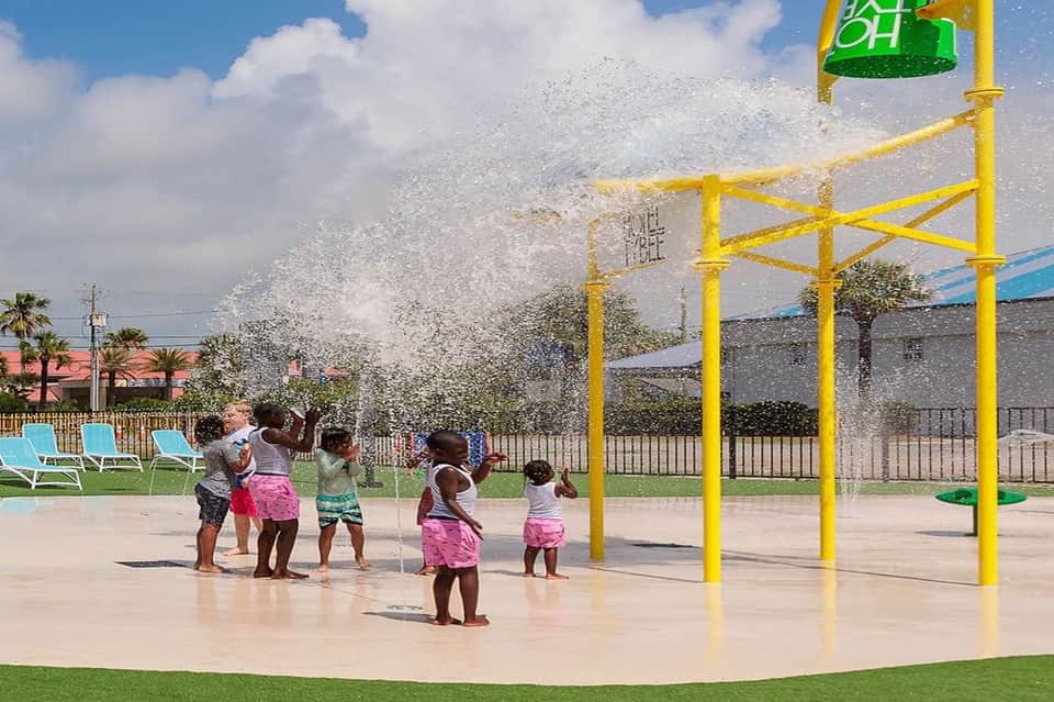 Children playing under yellow water spray structure with bucket dump feature on wet deck