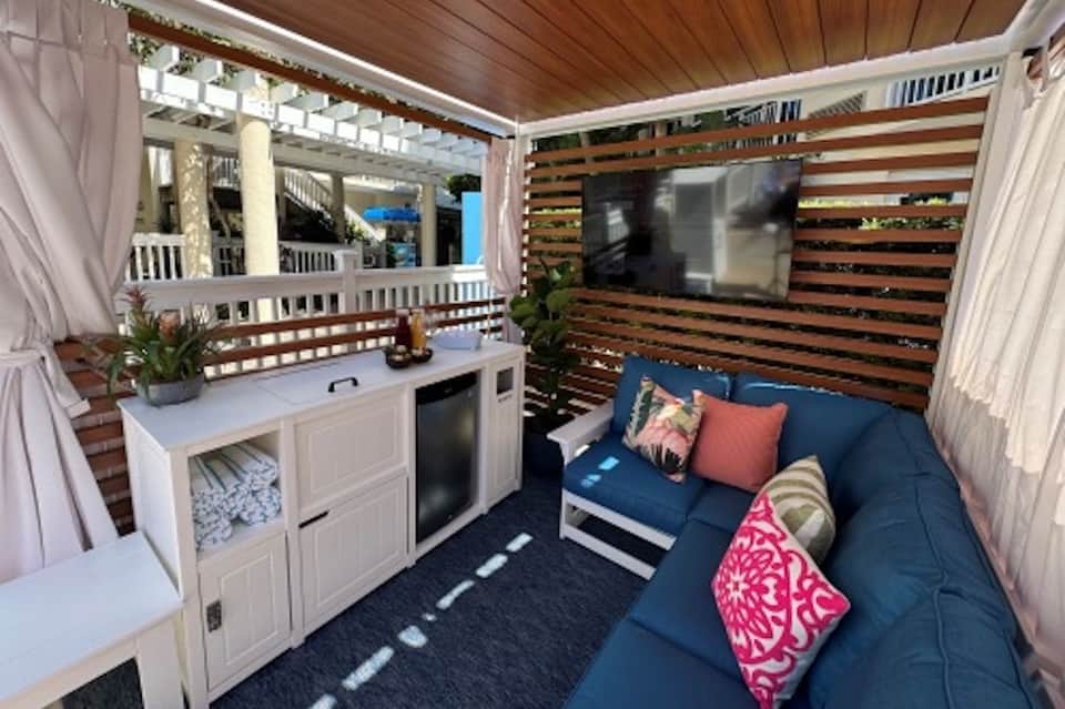 Modern beach cabana interior with blue sofa, kitchenette, wood ceiling, and entertainment screen