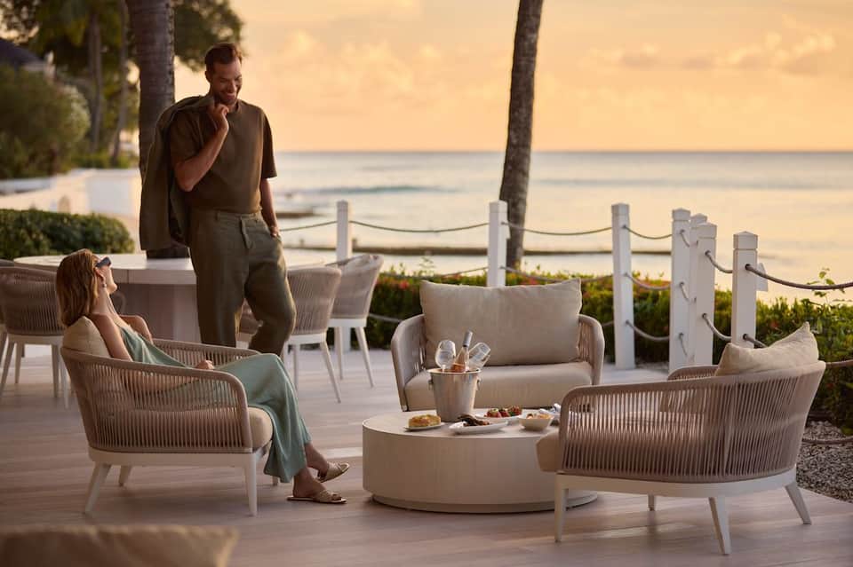 Couple relaxing on a beachfront deck with woven chairs, tropical plants, and calm ocean at dusk