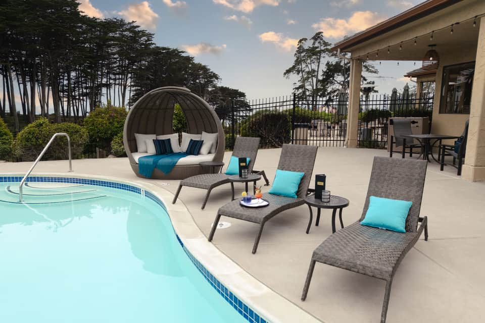 Pool deck with three gray chaise lounges, round ball chair, and covered patio area near crystal blue pool
