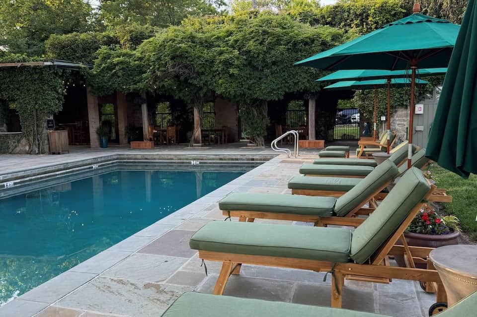 Resort pool with row of cushioned lounge chairs, green umbrellas, and ivy-covered building