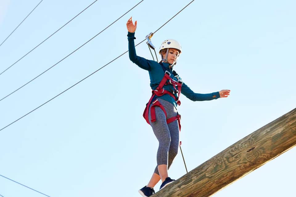 Woman zip-lining on wooden beam wearing safety helmet and harness with cables overhead