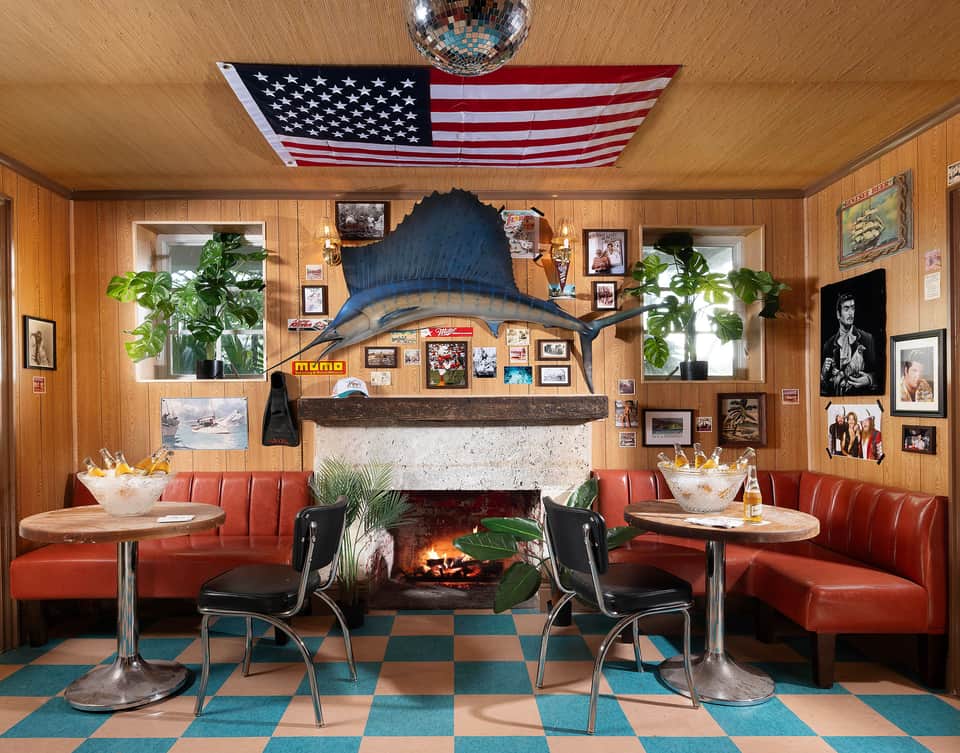 Vintage lodge interior with fireplace, sailfish mount, American flag, and red seating