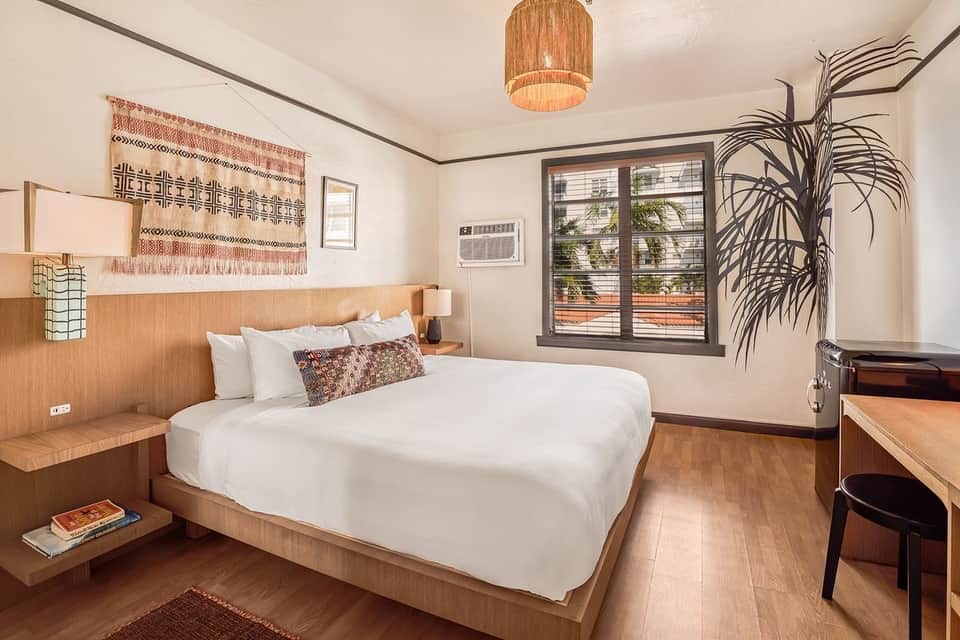 Modern hotel bedroom with wooden bed frame, white linens, woven wall hanging, potted plant, and large windows with views