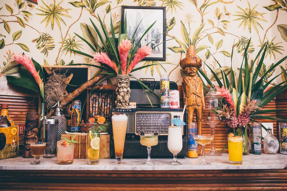 Tropical-themed bar with floral wallpaper, wooden carved figures, pink flowers, vintage radio, and colorful cocktails on counter