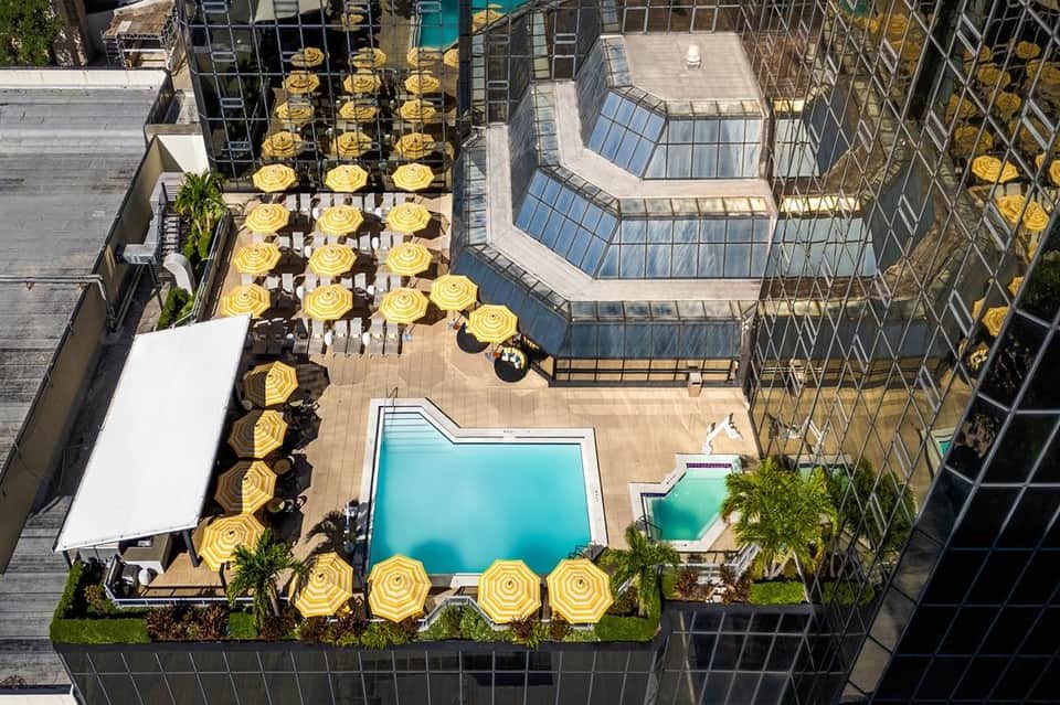 Top-down view of urban rooftop pool area with turquoise water, yellow umbrellas, lounge chairs, and tall buildings