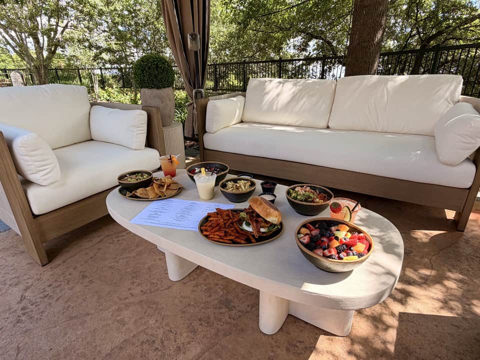 Upscale outdoor dining setup with cream sofa, armchair, food spread on white table, and wooded backdrop