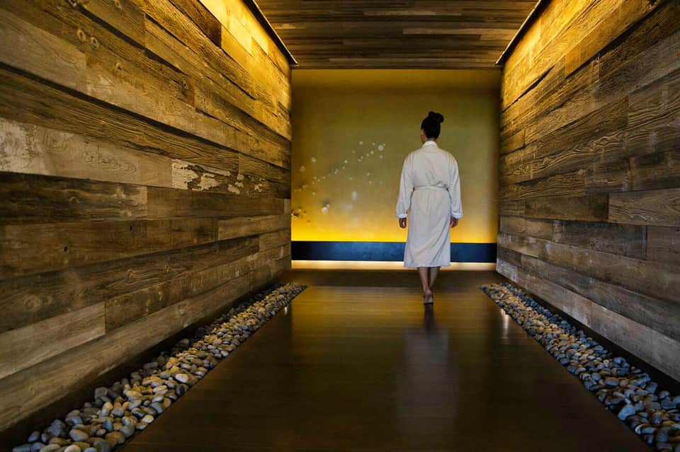 Woman in spa robe walking through modern wellness corridor with wooden walls and illuminated yellow accent lighting