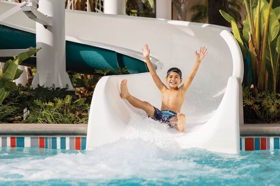 Child sliding down white water slide into bright turquoise pool with tropical plants on sides