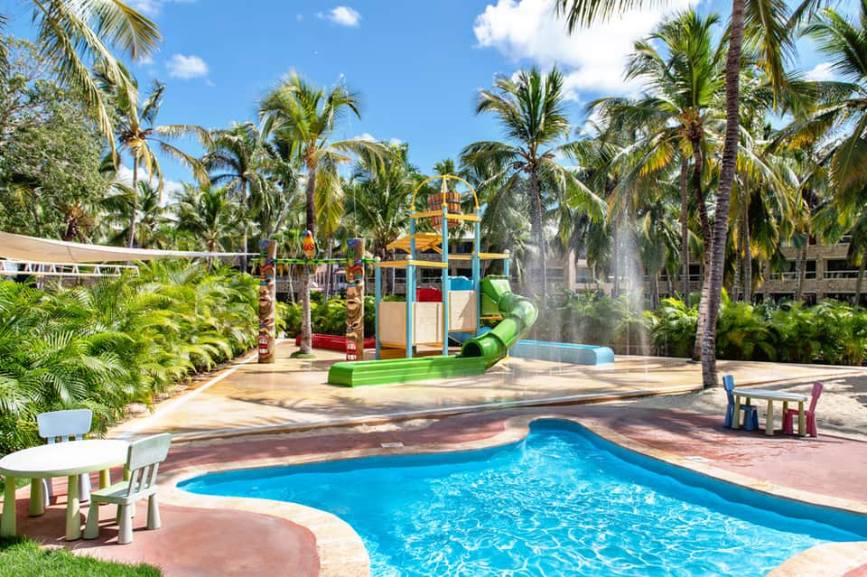 Resort pool with colorful playground, water slide, and palm trees in tropical setting