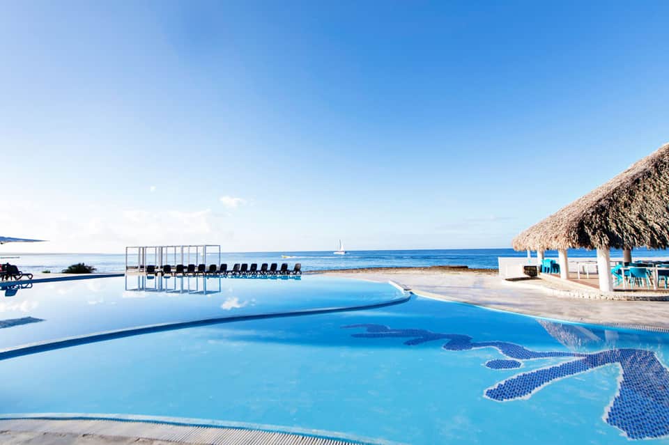 Curved infinity pool overlooking ocean beach with lounge chairs and thatched-roof cabana