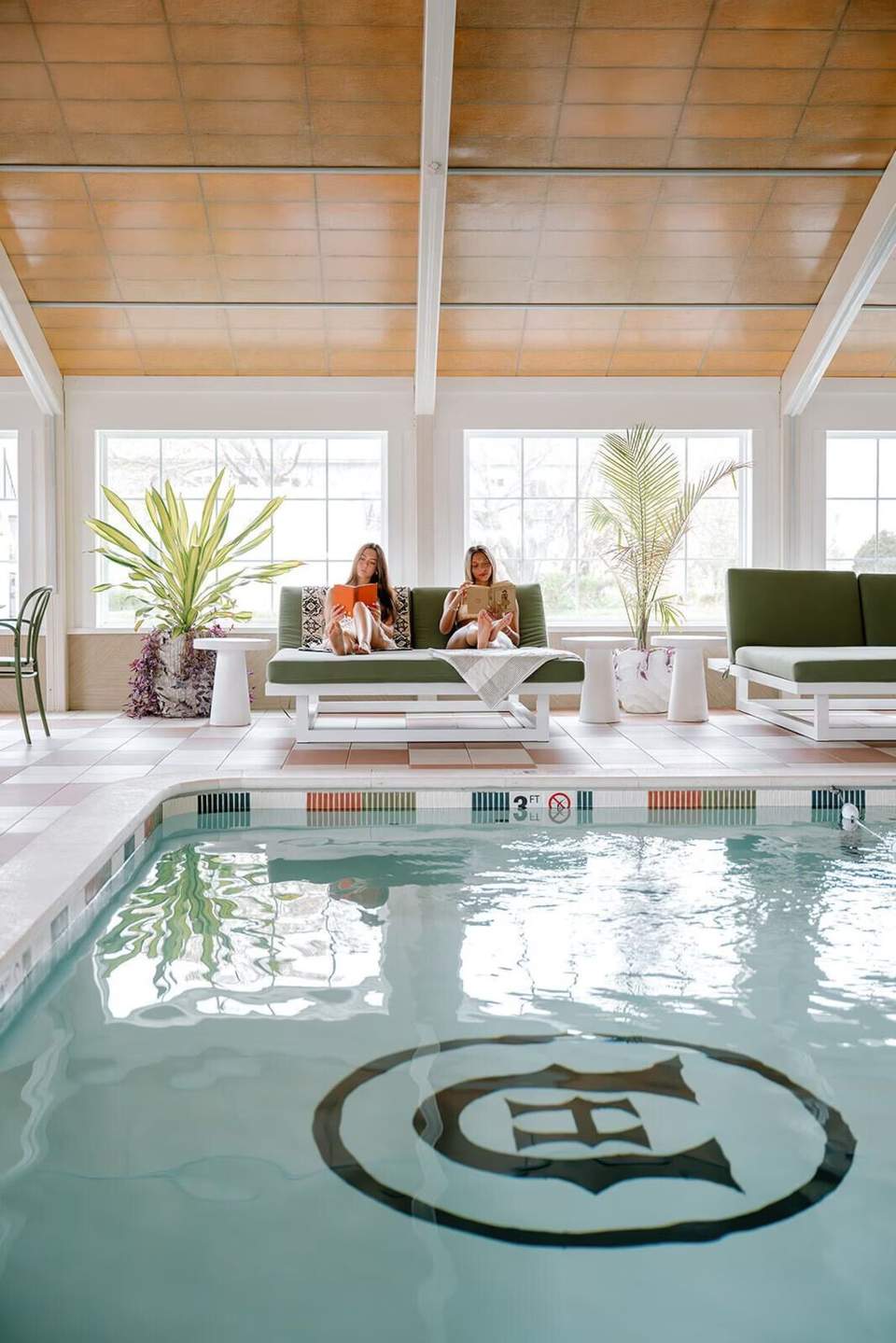 Indoor pool facility with wooden ceiling, large windows, green seating, potted plants, and natural light