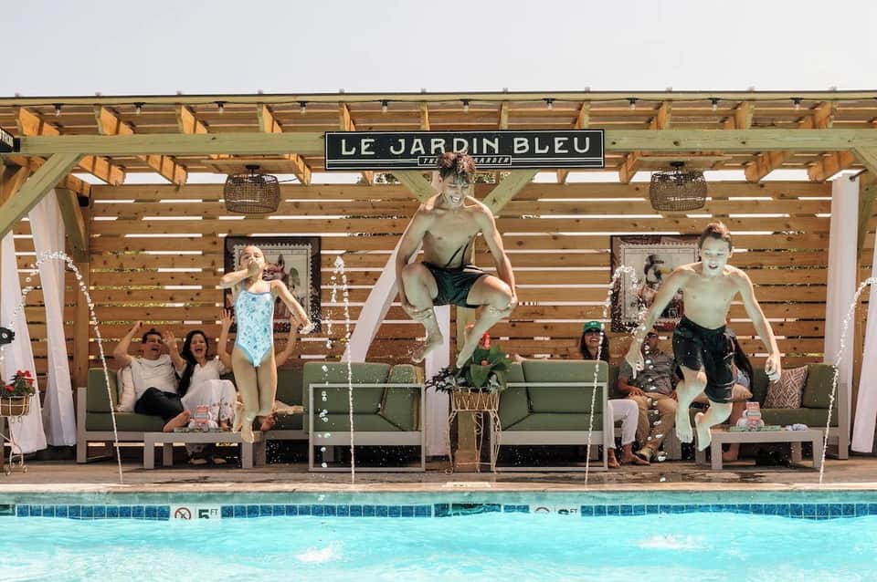 Poolside cabanas with vintage-style statues, green seating, white drapes, and wooden pergola structures