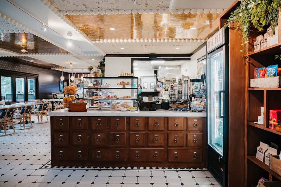 Modern bakery/café interior with wooden display drawers, glass cases, white ceiling details, and product shelving
