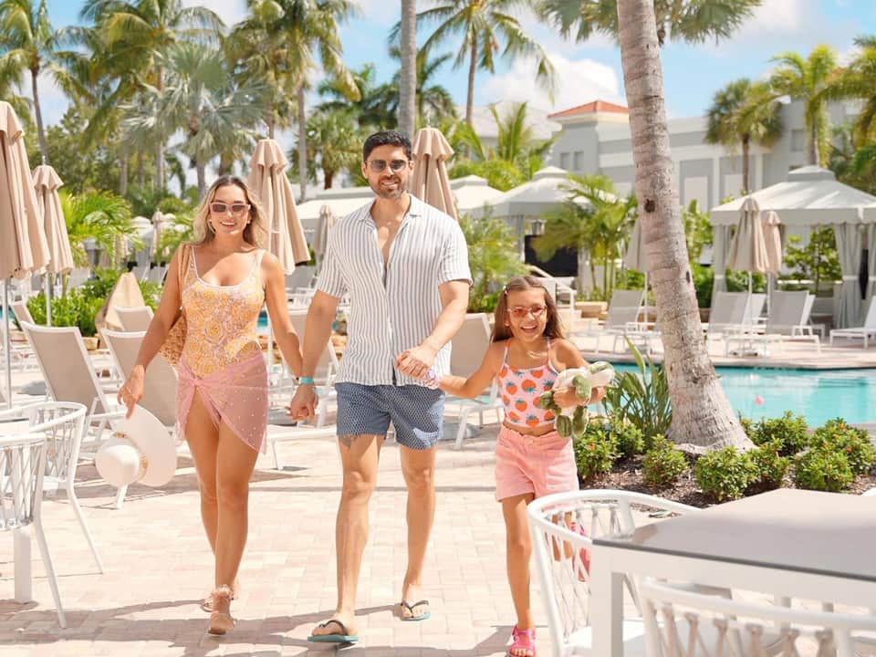 Family walking on resort pool deck with white lounge chairs, umbrellas, and palm trees