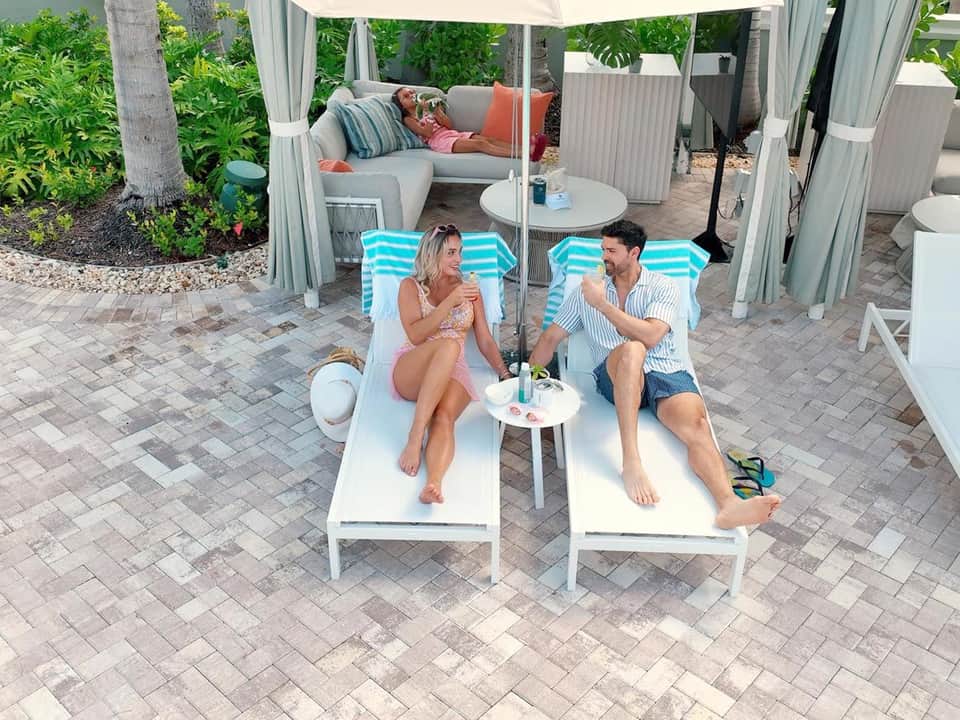 Couple relaxing on striped lounge chairs under white canopy tent with coastal greenery backdrop