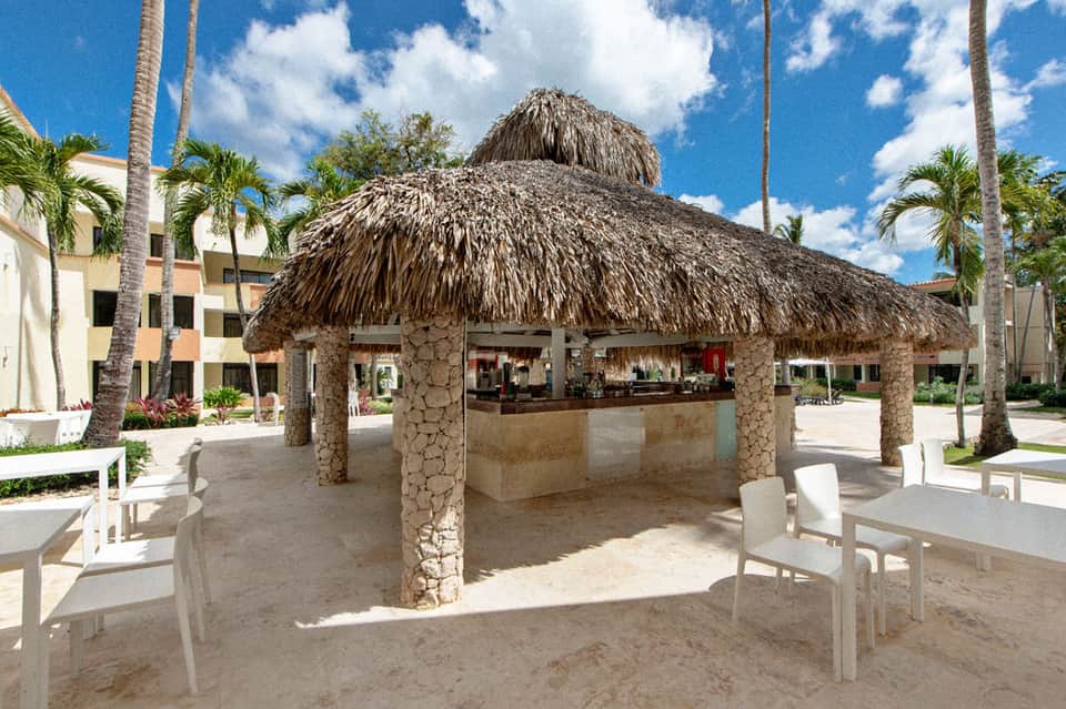 Thatched-roof beach bar with stone columns, palm trees, and white seating on sandy grounds