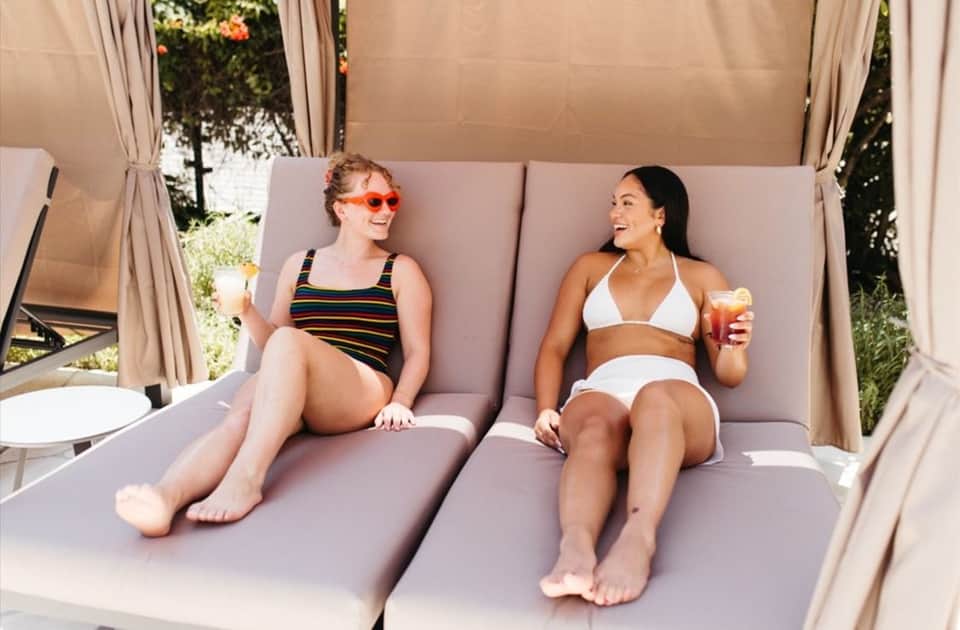 Two women in bikinis sitting on purple cushioned daybed with beige canopy curtains and garden view