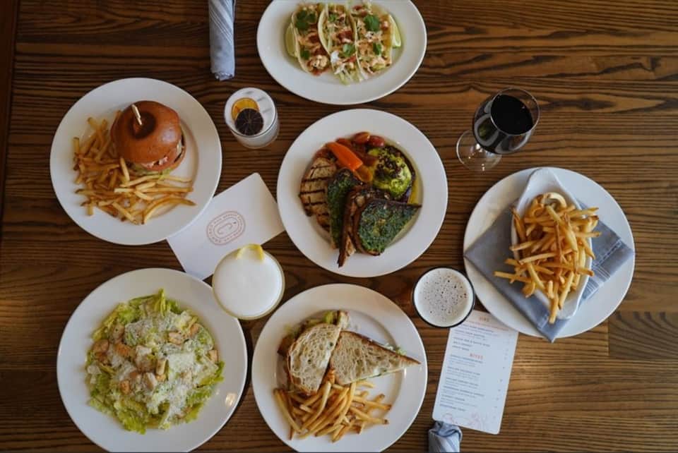 Overhead view of multiple plates with burgers, tacos, grilled fish, salads, fries, and beverages on wooden table