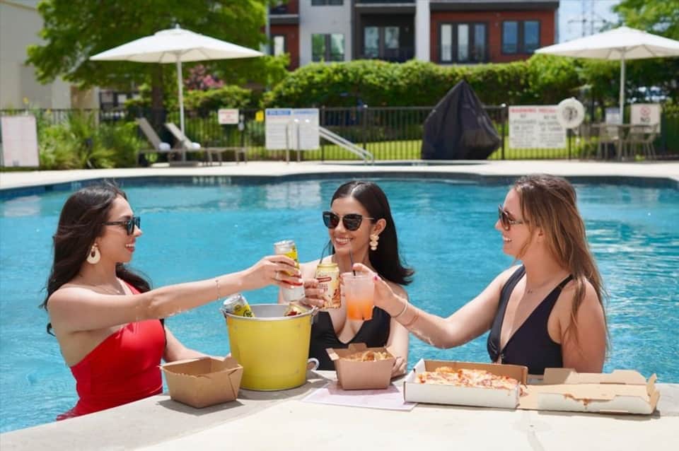 Three women toasting with drinks while sitting in pool at resort with umbrellas and landscaped grounds