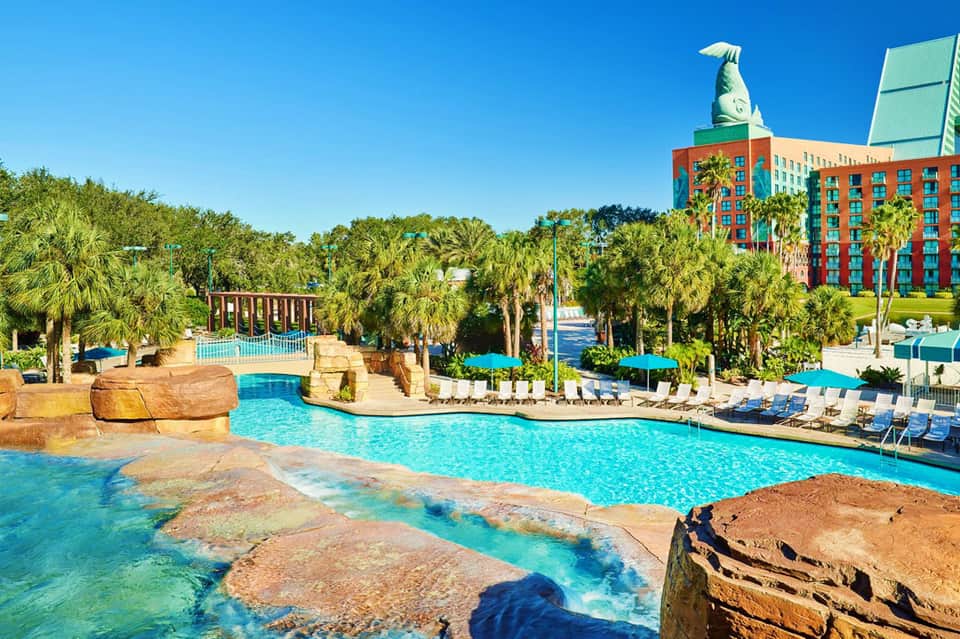 Resort pool with turquoise water, palm trees, and colorful orange building with large fish sculpture on roof
