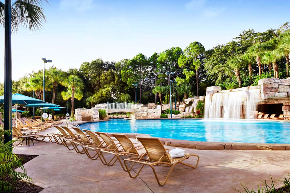 Resort pool with waterfall features, lounge chairs, palm trees, and tropical landscaping under blue sky