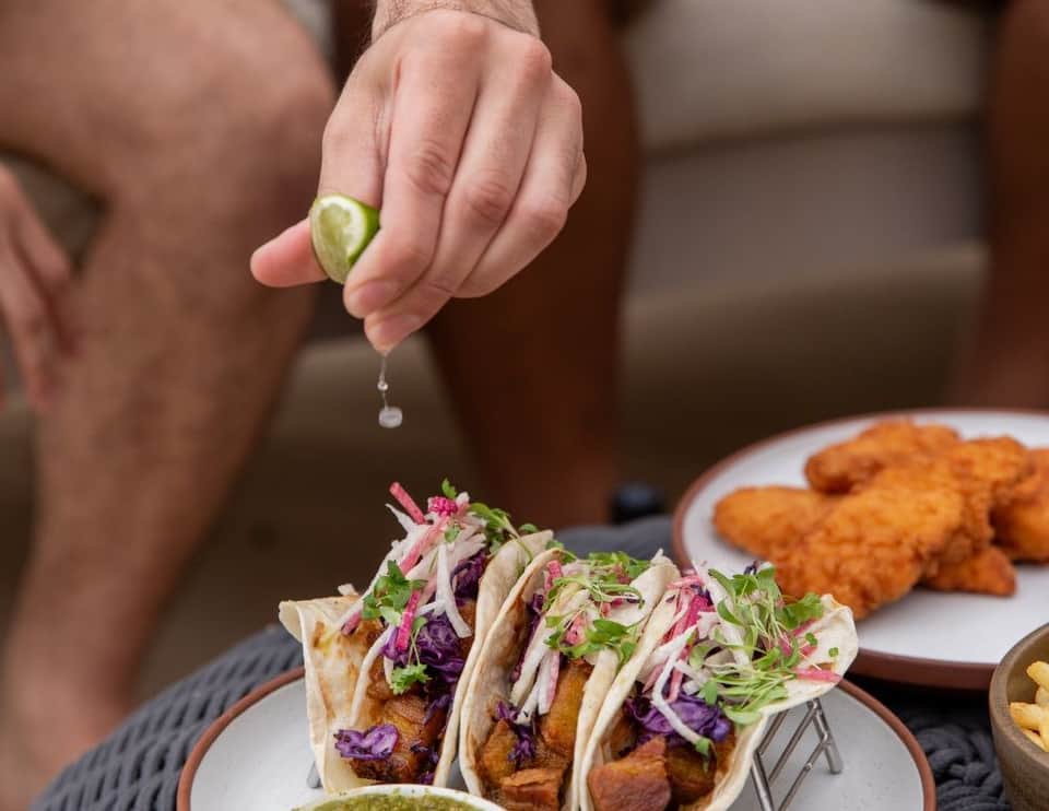 Hand squeezing lime over fish tacos with purple cabbage slaw and fried chicken wings on plates