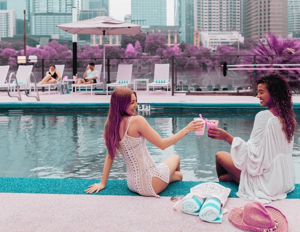 Two women in summer dresses toasting drinks by a pool with purple flowering trees and city skyline backdrop