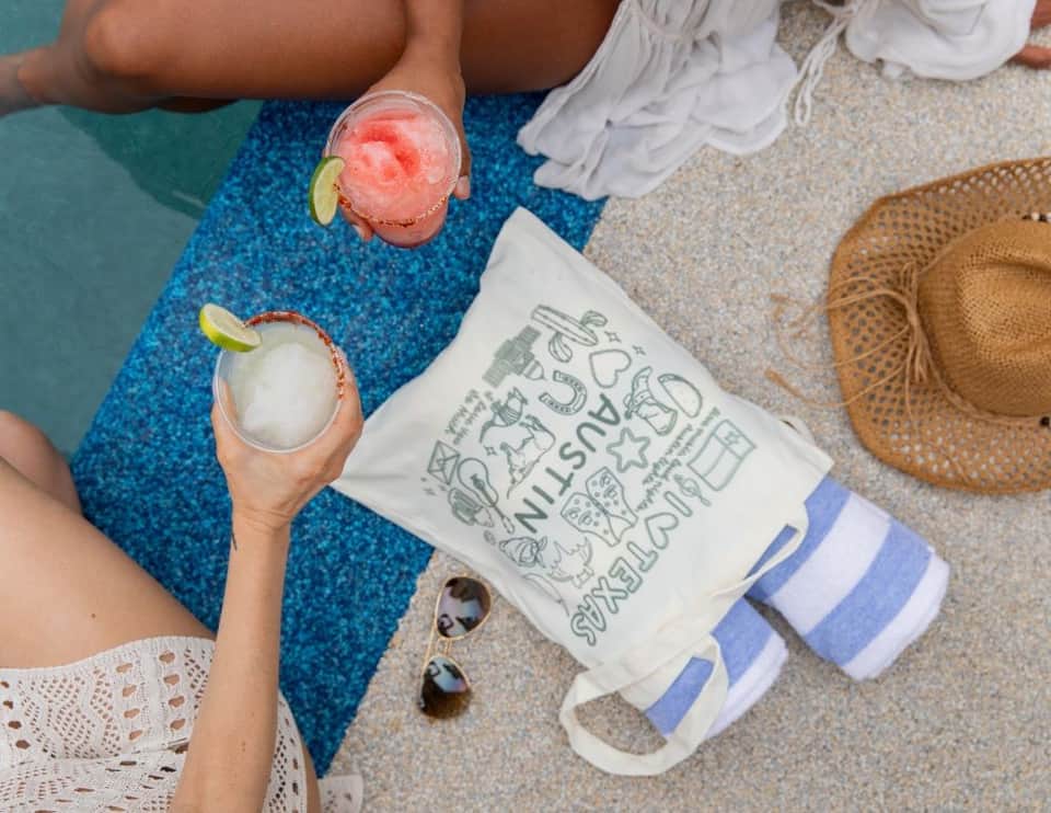 Overhead view of two colorful cocktails with lime garnish held by someone poolside on a striped towel