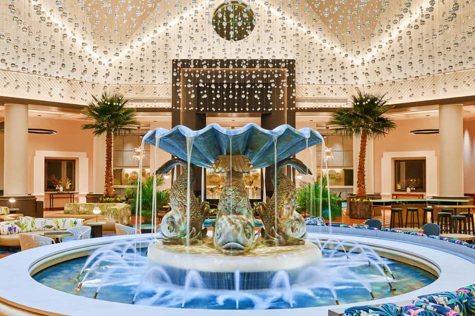 Luxurious resort atrium with circular fountain, crystal chandelier ceiling, and palm trees