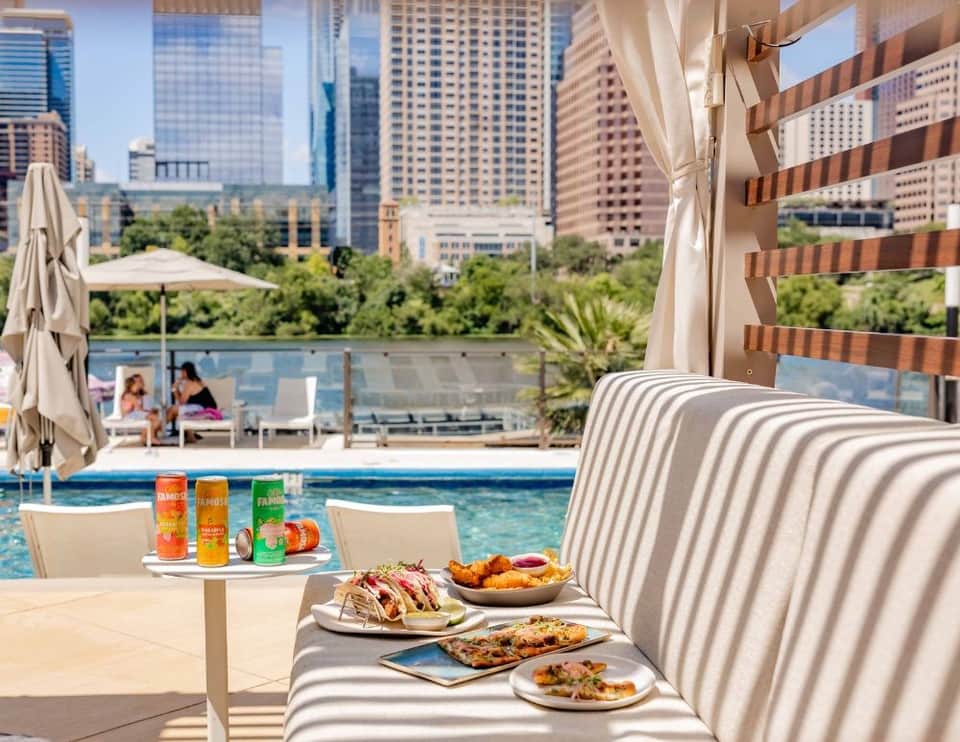 Waterfront pool area with city skyline views, striped lounge chairs, umbrellas, and gourmet food service