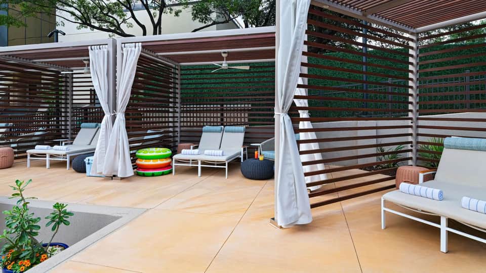 Modern resort deck with lounge chairs, white curtains, wooden pergolas, and colorful pool floats