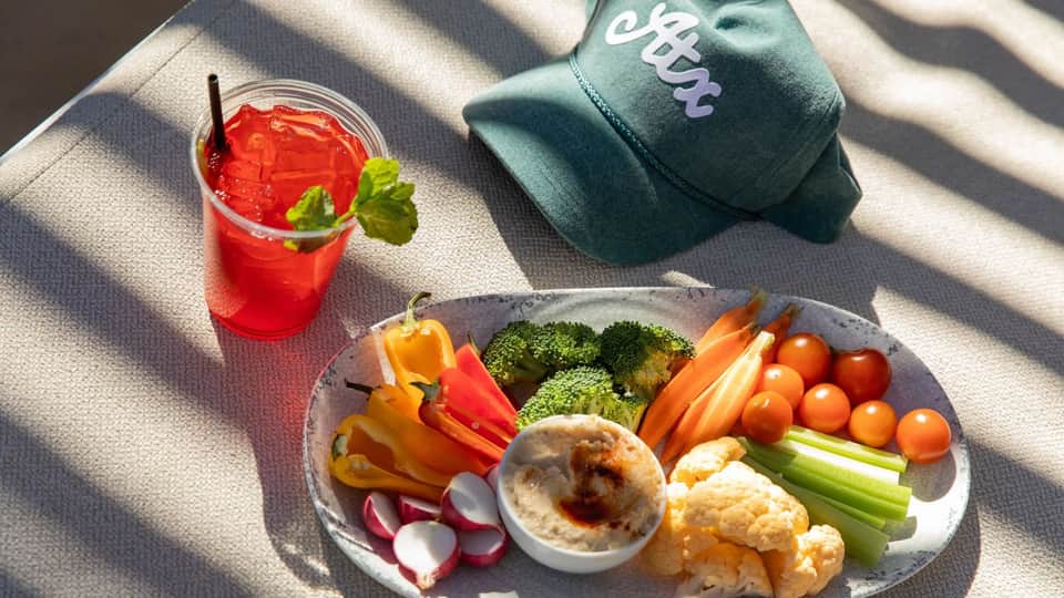 Healthy snack spread on lounge chair with fresh vegetables, hummus, and red beverage