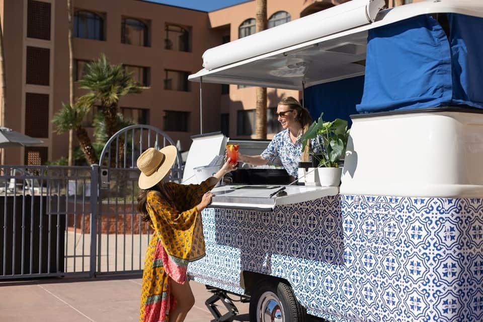 Woman ordering from colorful food cart at resort courtyard with palm trees and buildings