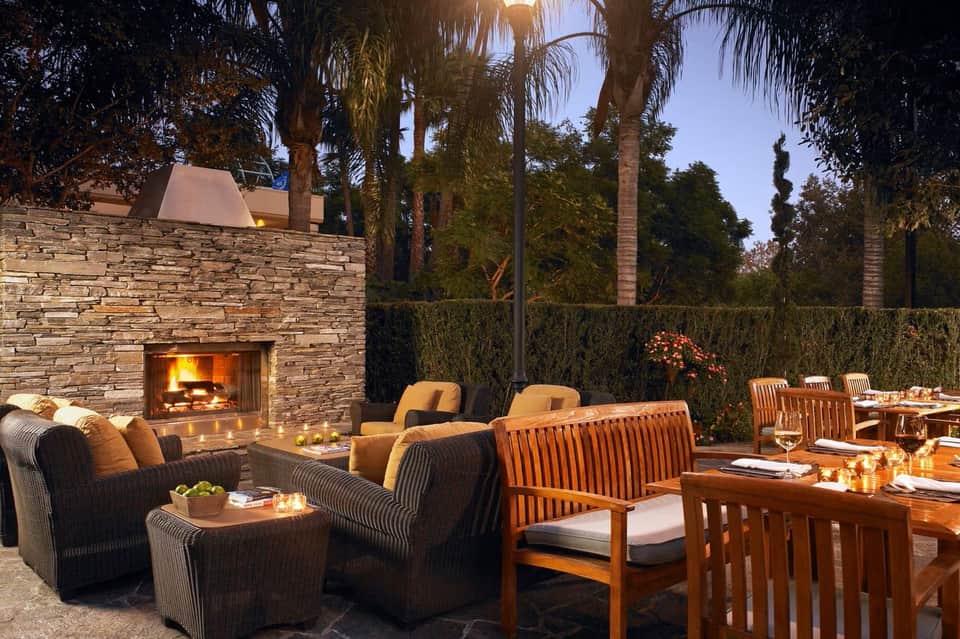 Outdoor fireplace lounge at dusk with stone walls, wooden furniture, and palm trees