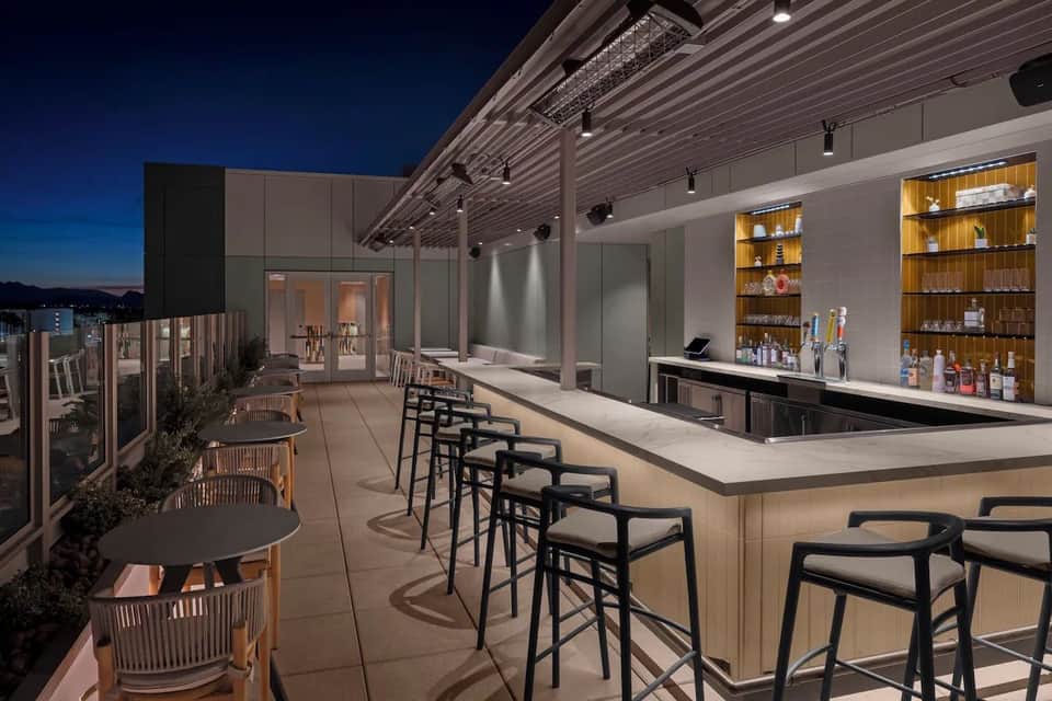 Rooftop bar at night with wooden shelving, white counter, and black bar seating